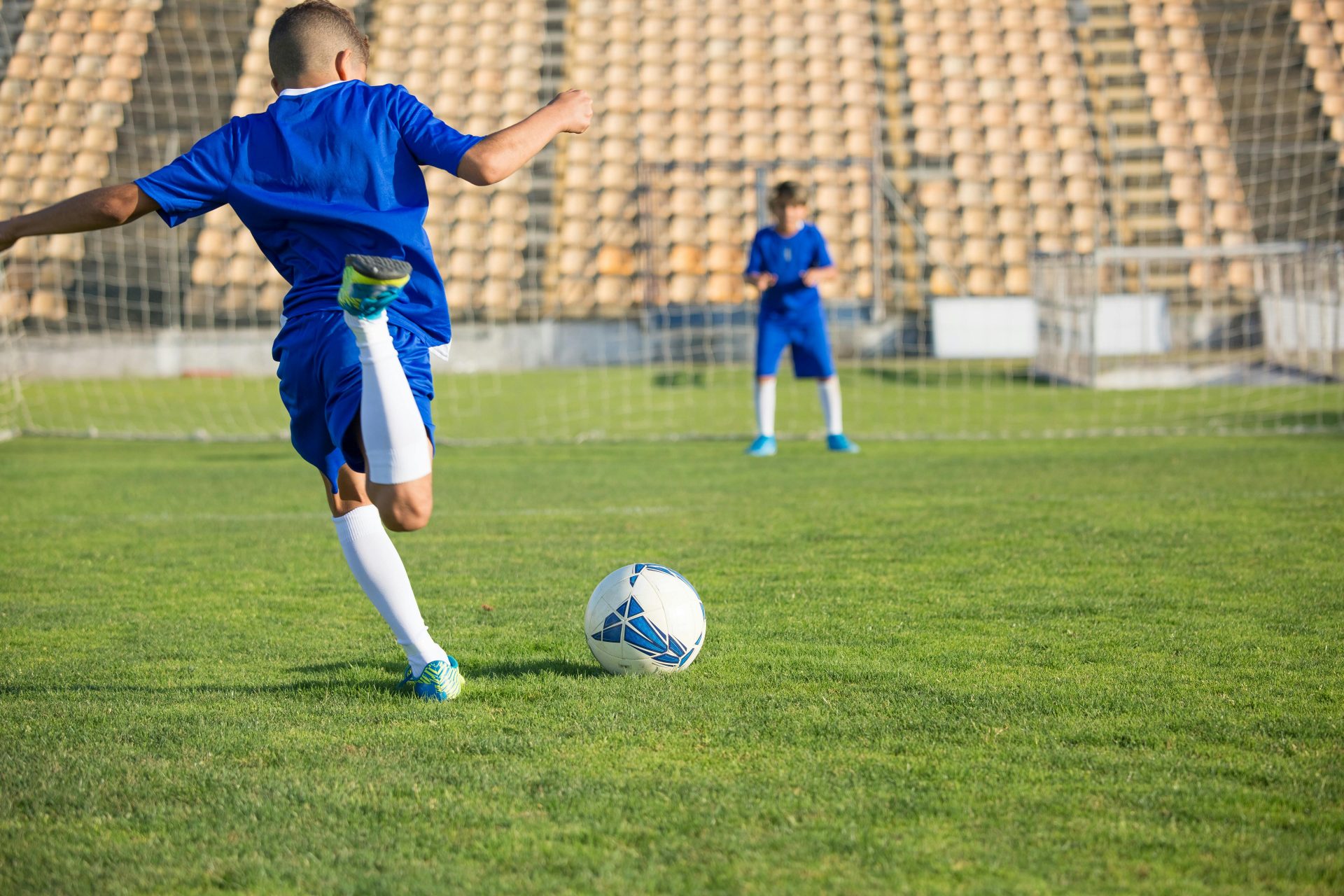 Child dressed in blue jersey and shorts about to kick a soccer ball to his friend.